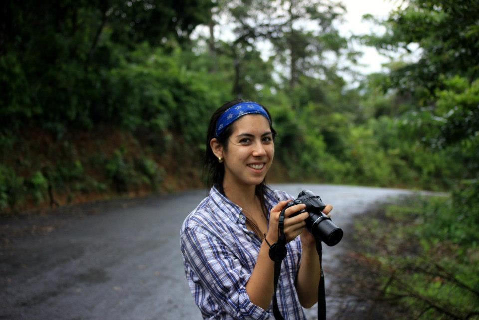 Anna Kudla stands in the foreground, smiling toward the camera. She is wearing a plaid shirt and blue bandana, holding a camera at the ready in front of her. Behind her, out of focus, a road curves into the lush rainforest.