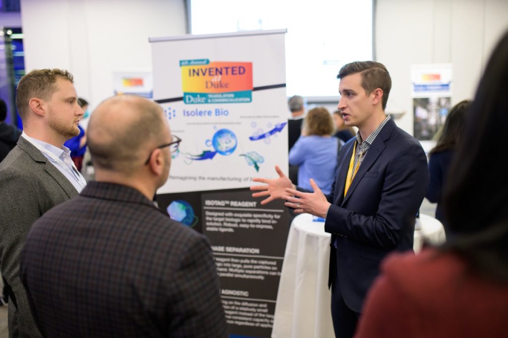 Michael Dzuricky stands to the right of the photo, actively gesturing with his hands as he describes Isolere Bio's technology to two attendees of Invented at Duke. People are wearing suits and in the background, slightly out of focus, is a cocktail table with white linen and a poster featuring graphics and text about Isolere Bio. Further back out of focus are glimpses of a crowd of attendees and other posters.