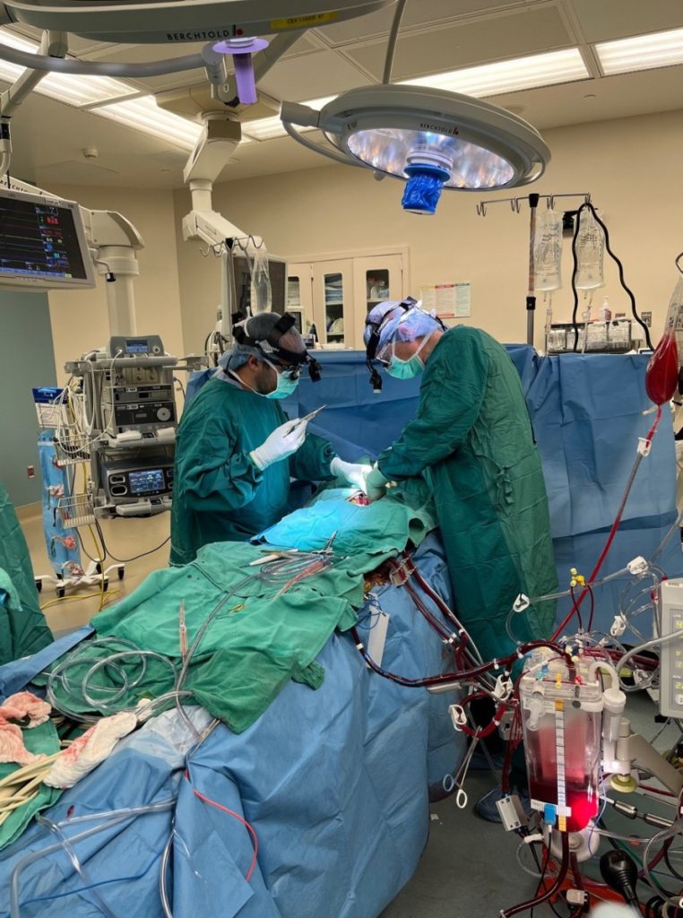 In an operating room cluttered with tubes and equipment, two surgeons fully geared up in green scrubs, masks, and gloves lean over a patient who is covered with blue and green surgical sheets.