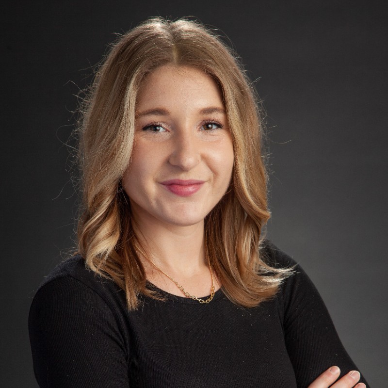 Headshot of Cambre Kelly in a black shirt, arms crossed out of frame, smiling at the camera, on a grey background.