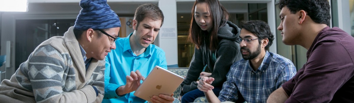 duke engineering entrepreneurship background students and professor huddled around working on an ipad