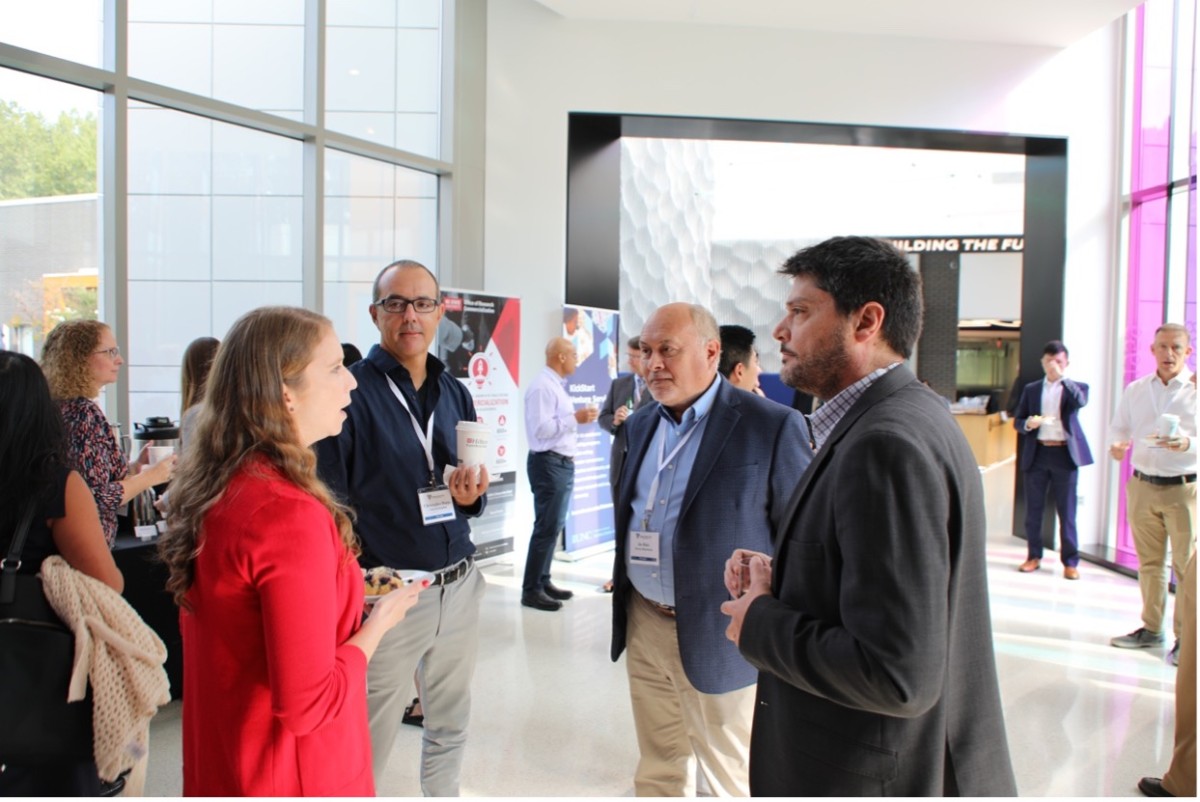 In the foreground, a group of four people in business attire chat in an airy white atrium filled with light streaming in from large windows. Around them and in the background, a few more people in business attire are milling about with coffee cups.