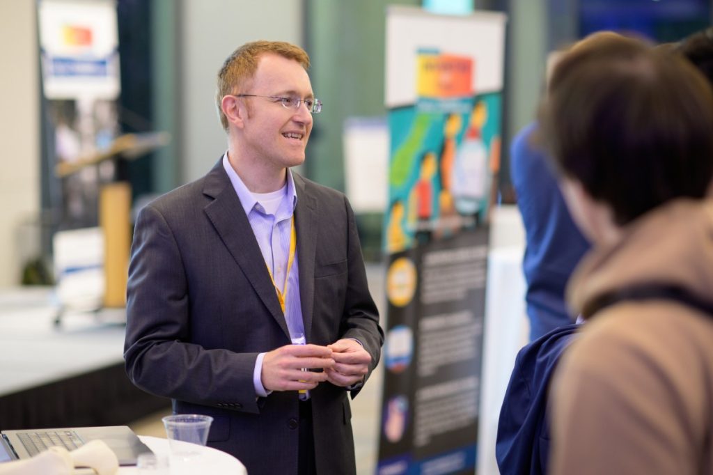 Benjamin Wiley smiles in a suit standing next to a cocktail table in a mingling setting, talking to out-of-frame attendees of Invented at Duke 2022.