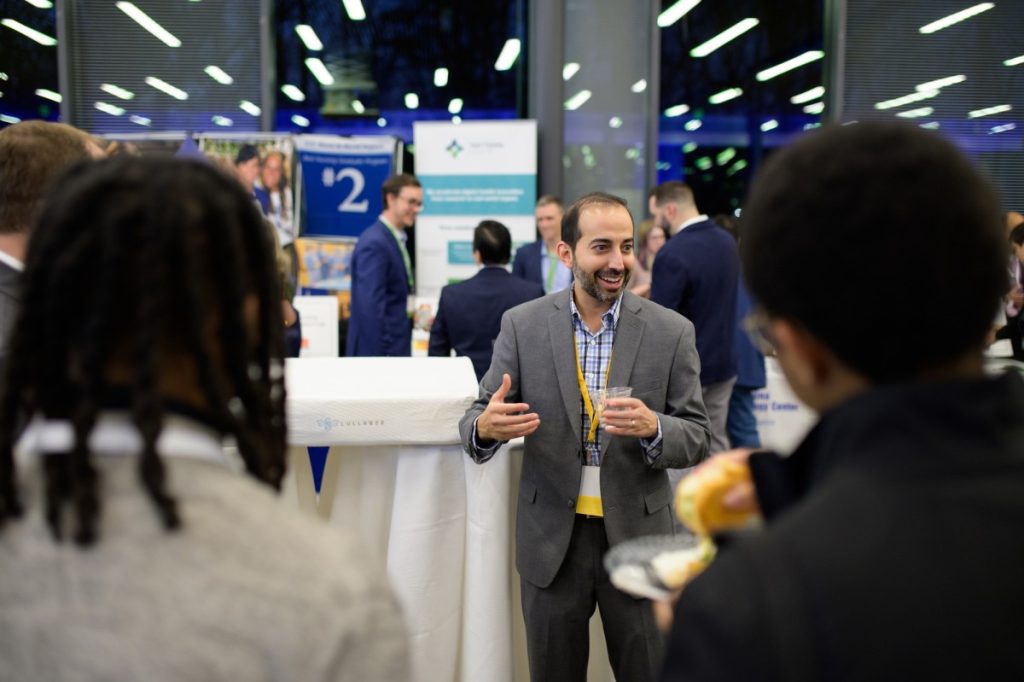 Sujay stands facing the camera and a group of out-of-focus attendees in the foreground, smiling and gesturing. A small white mattress lays on a cocktail table beside him, emblazoned with the Lullabee logo.