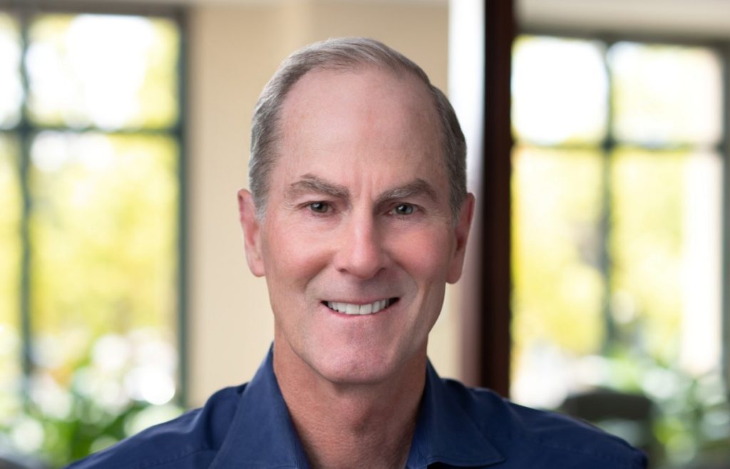 Ned Gilhuly headshot, smiling at the camera in an out-of-focus room with greenery shining through the windows behind him.