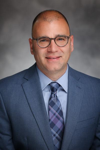 Headshot of Justin Pollara, smiling at the camera in a grey-blue suit.