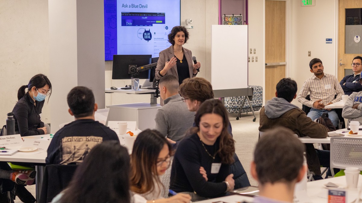 Jones stands in the front of a room with a microphone, speaking to multiple groups of professional students seated around triangular tables.