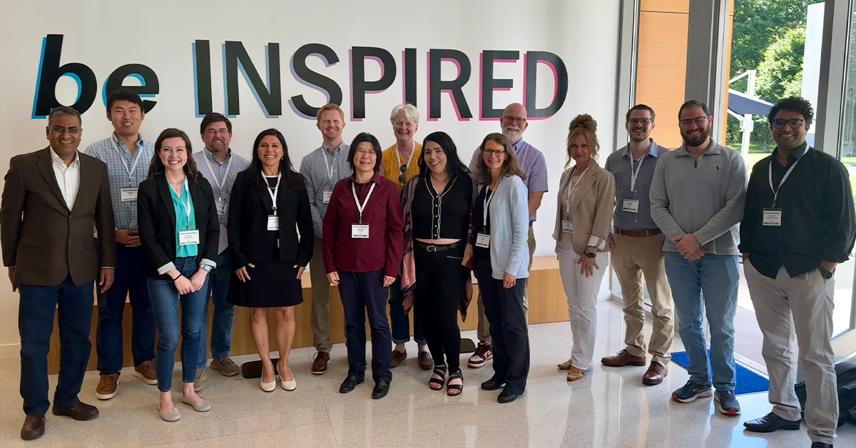 Fifteen people in business casual stand in a modern, light filled lobby smiling at the camera. Behind them, the words “be INSPIRED” are stenciled on the wall.