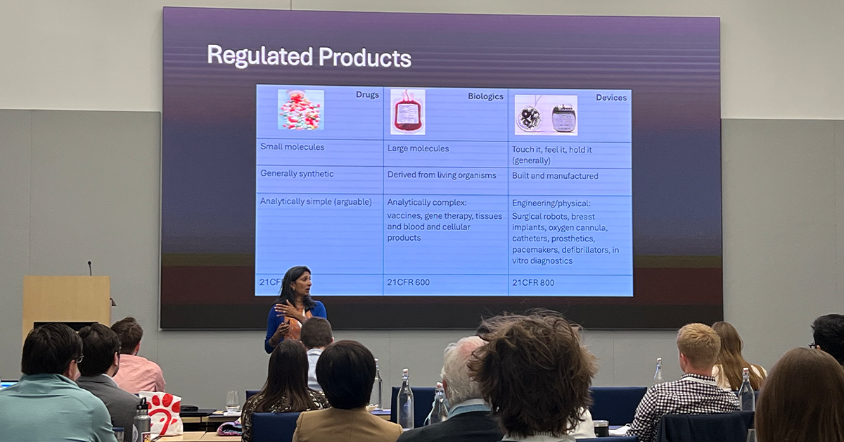 Speaker Raman stands at the front of a grey room with a large screen behind her which features a table of differences between FDA regulations of Drugs, Biologics, and Devices. Participants sit at long tables looking forward attentively.