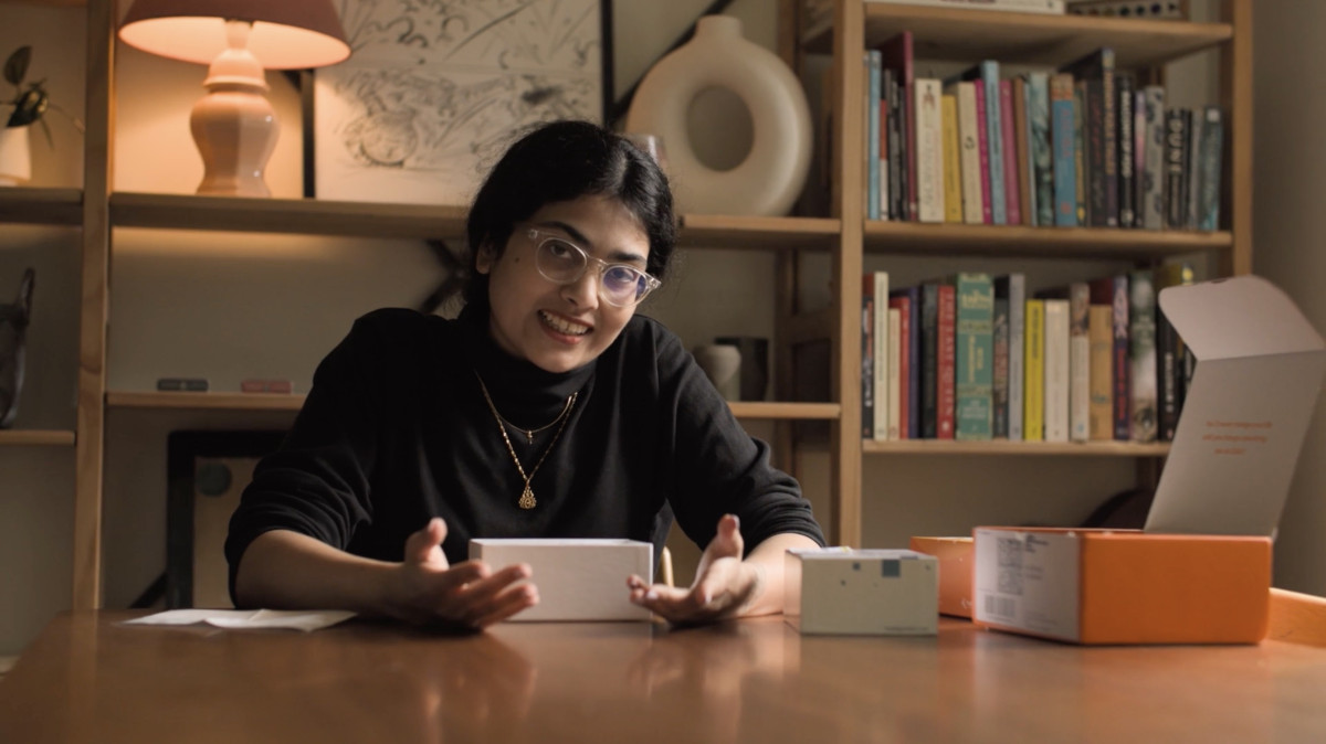 Ankita sits at a table with a few tissue-box sized cardboard boxes of the TruAge Test kit. She’s gesturing with her hands open palms and forward around one of the boxes. The room is warm, lots of wood, with a tidy bookshelf behind her.