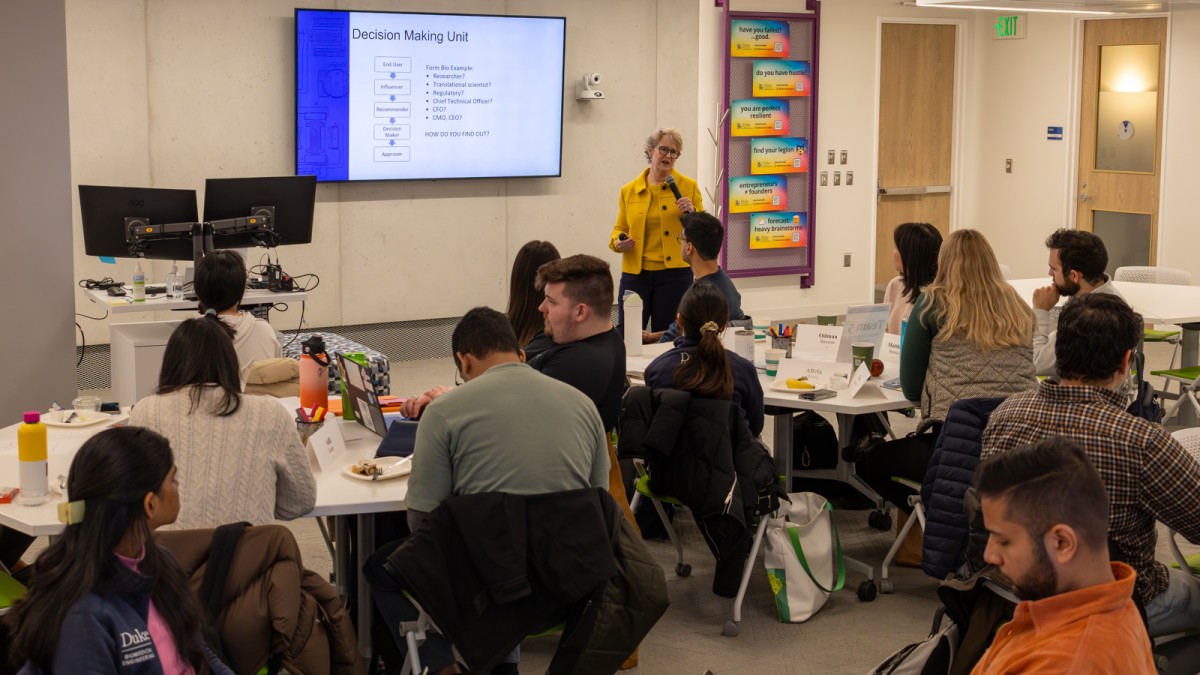 About a dozen students sit around several tables in a classroom workshop environment, looking toward the front of the room, where Claire Aldridge is standing with a handheld microphone mid-lecture.