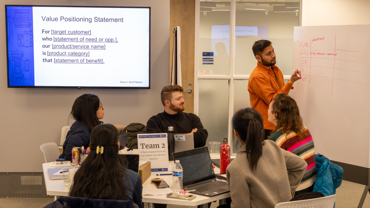 Workshop classroom environment, with 5 students seated around a table, looking to a standing Yash Shah writing on a roller whiteboard.